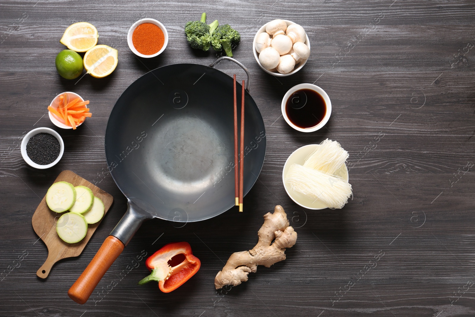 Photo of Empty iron wok and chopsticks surrounded by ingredients on dark grey wooden table, flat lay. Space for text