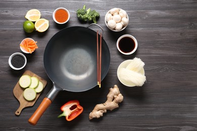 Photo of Empty iron wok and chopsticks surrounded by ingredients on dark grey wooden table, flat lay. Space for text