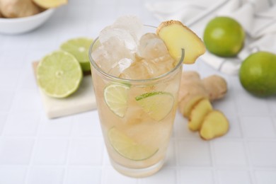 Photo of Glass of tasty ginger ale with ice cubes and ingredients on white tiled table, closeup