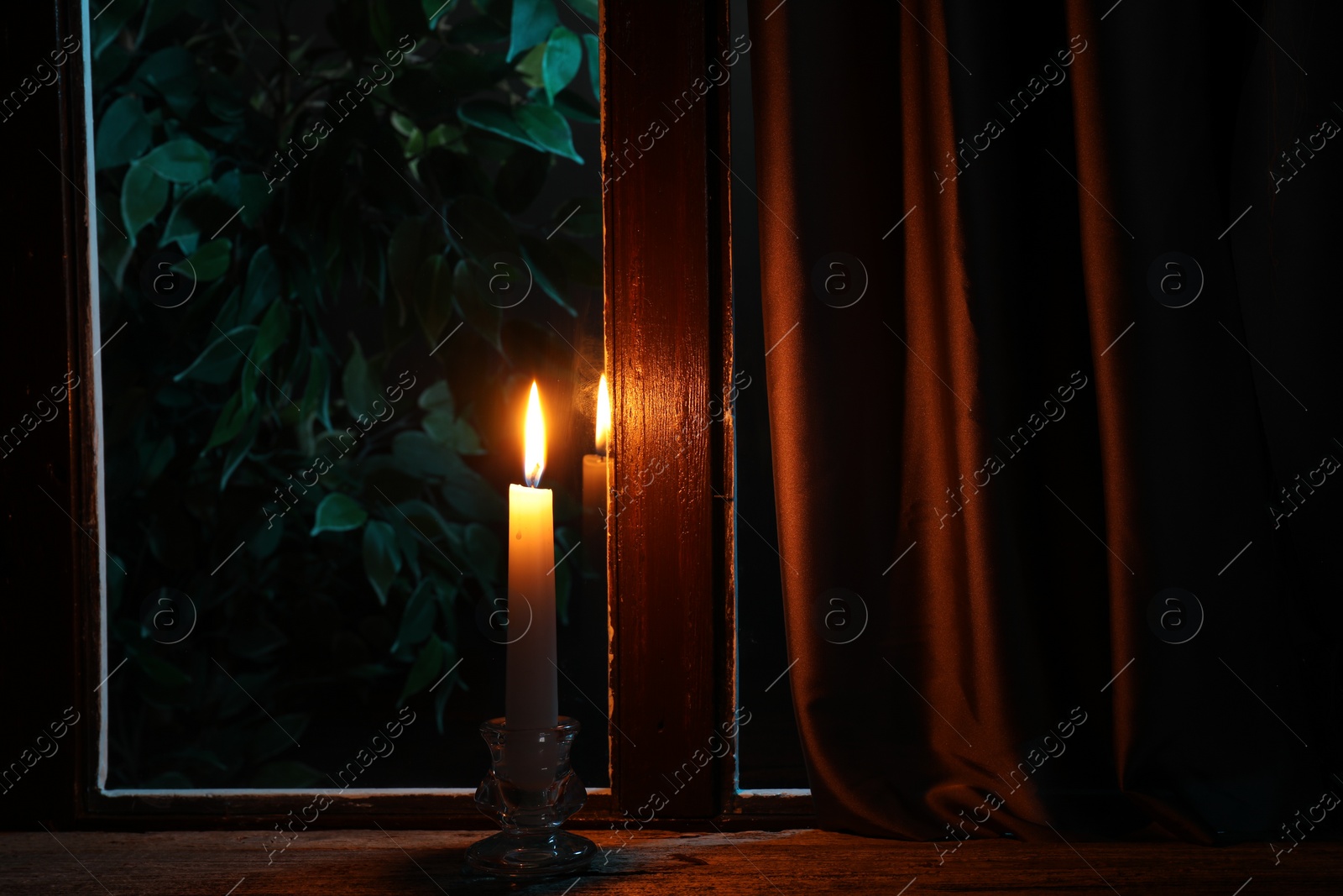 Photo of Burning candle on wooden table near window at night