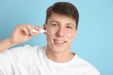 Teen guy with acne problem applying cream on light blue background