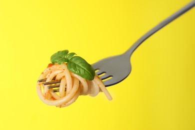 Fork with tasty pasta, basil and tomato sauce on yellow background, closeup