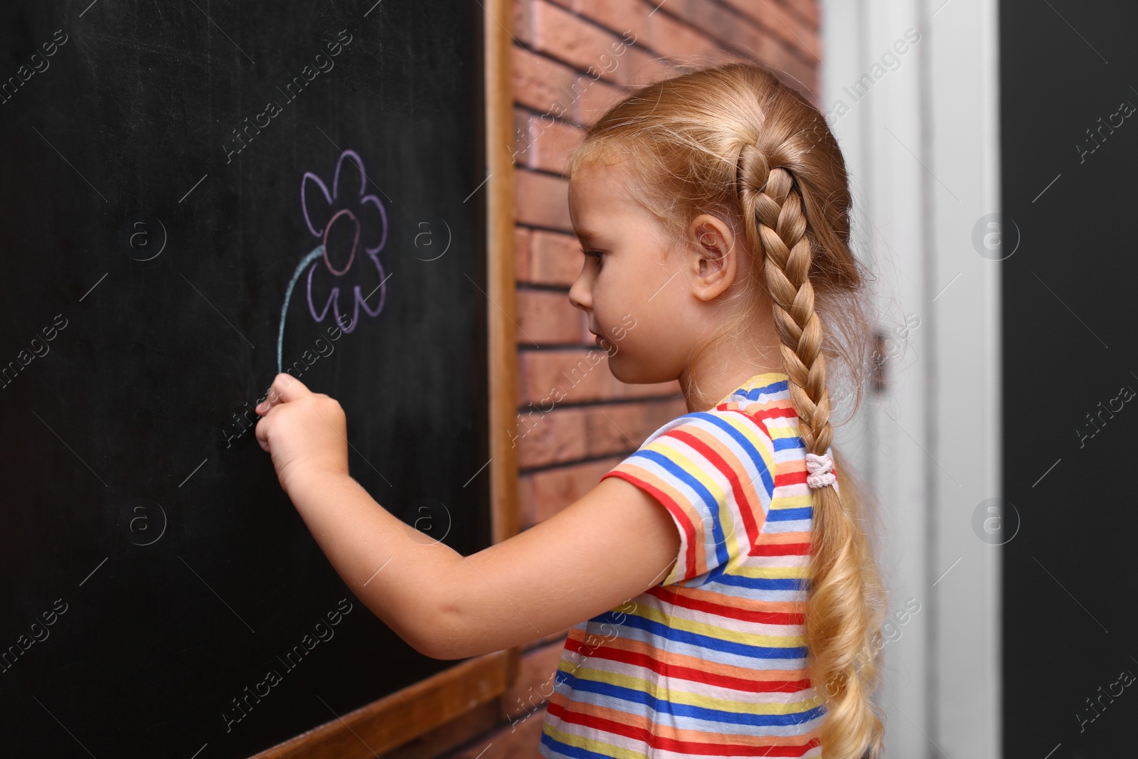 Photo of Cute little left-handed girl drawing on chalkboard near brick wall