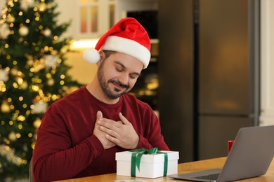 Photo of Celebrating Christmas online with exchanged by mail presents. Happy man in Santa hat thanking for gift during video call on laptop at home