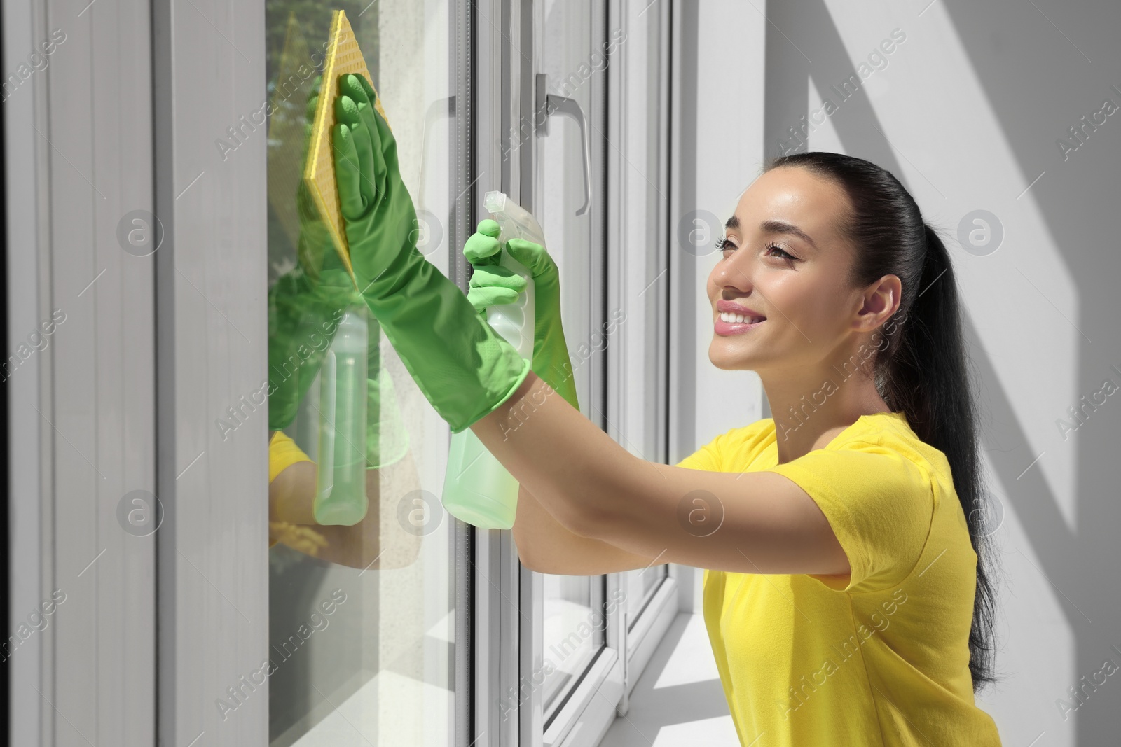 Photo of Happy young woman cleaning window glass with sponge cloth and spray indoors