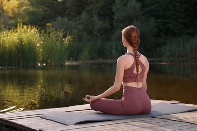 Photo of Woman practicing Padmasana on yoga mat on wooden pier near pond, back view. Lotus pose