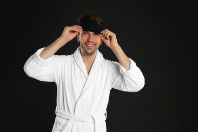 Photo of Happy young man in bathrobe and eye sleeping mask on black background