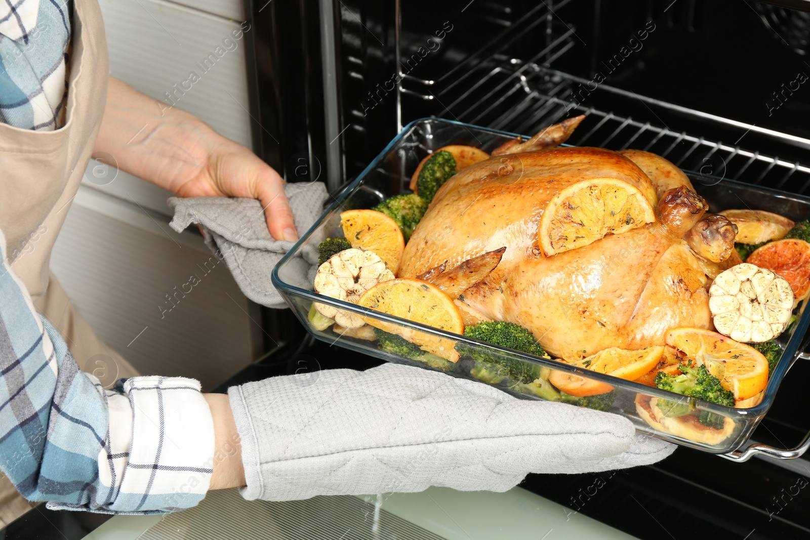 Photo of Woman taking baked chicken with oranges and vegetables out of oven, closeup