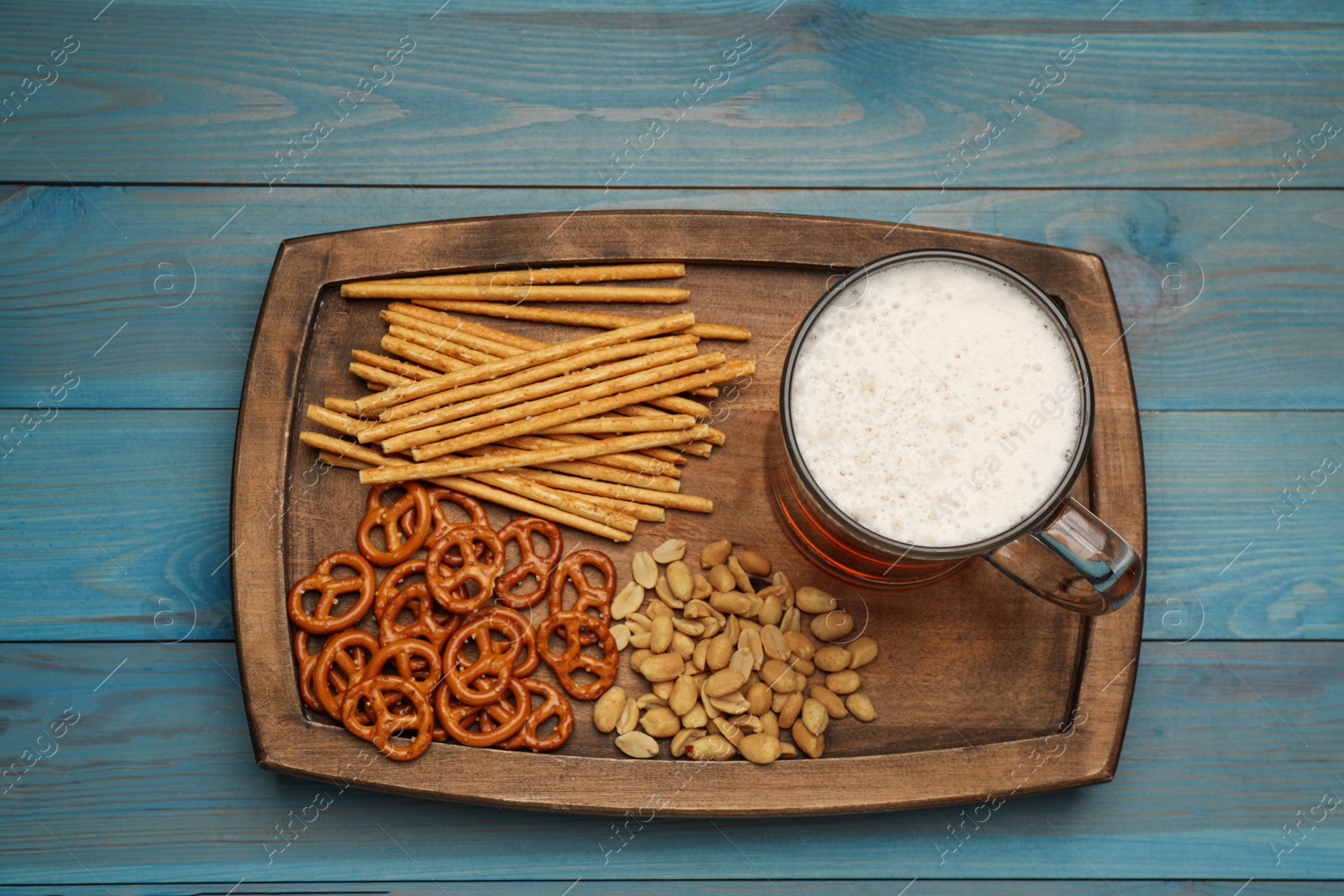 Photo of Delicious pretzel crackers, salted sticks, peanuts and mug of beer on light blue wooden table, top view