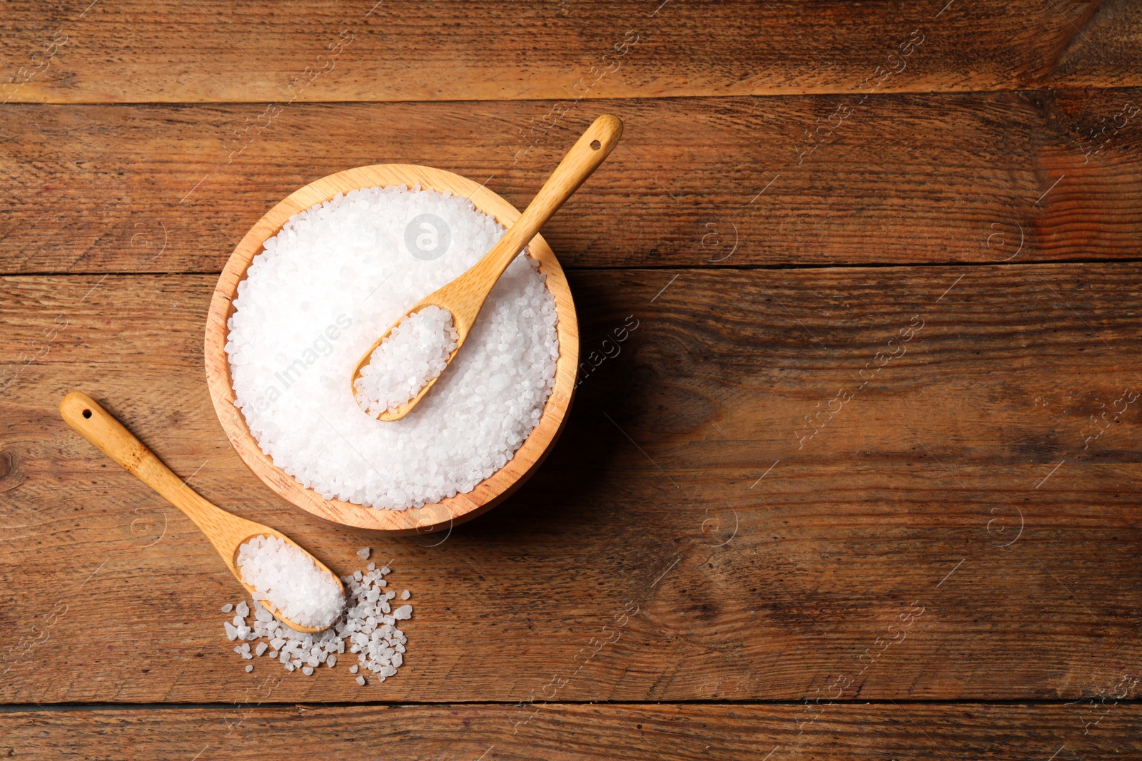 Photo of Spoons and bowl of natural sea salt on wooden table, flat lay. Space for text