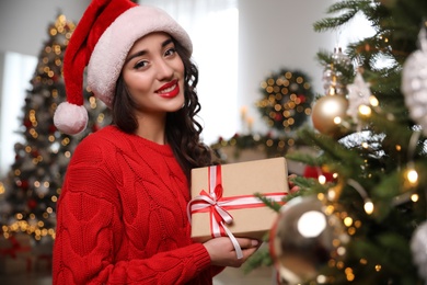 Photo of Beautiful woman in Santa hat with gift box near Christmas tree at home