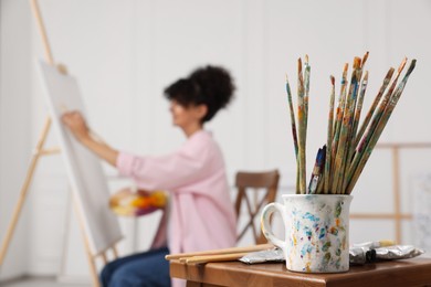 Photo of Young woman painting on easel with canvas in studio, focus on brushes