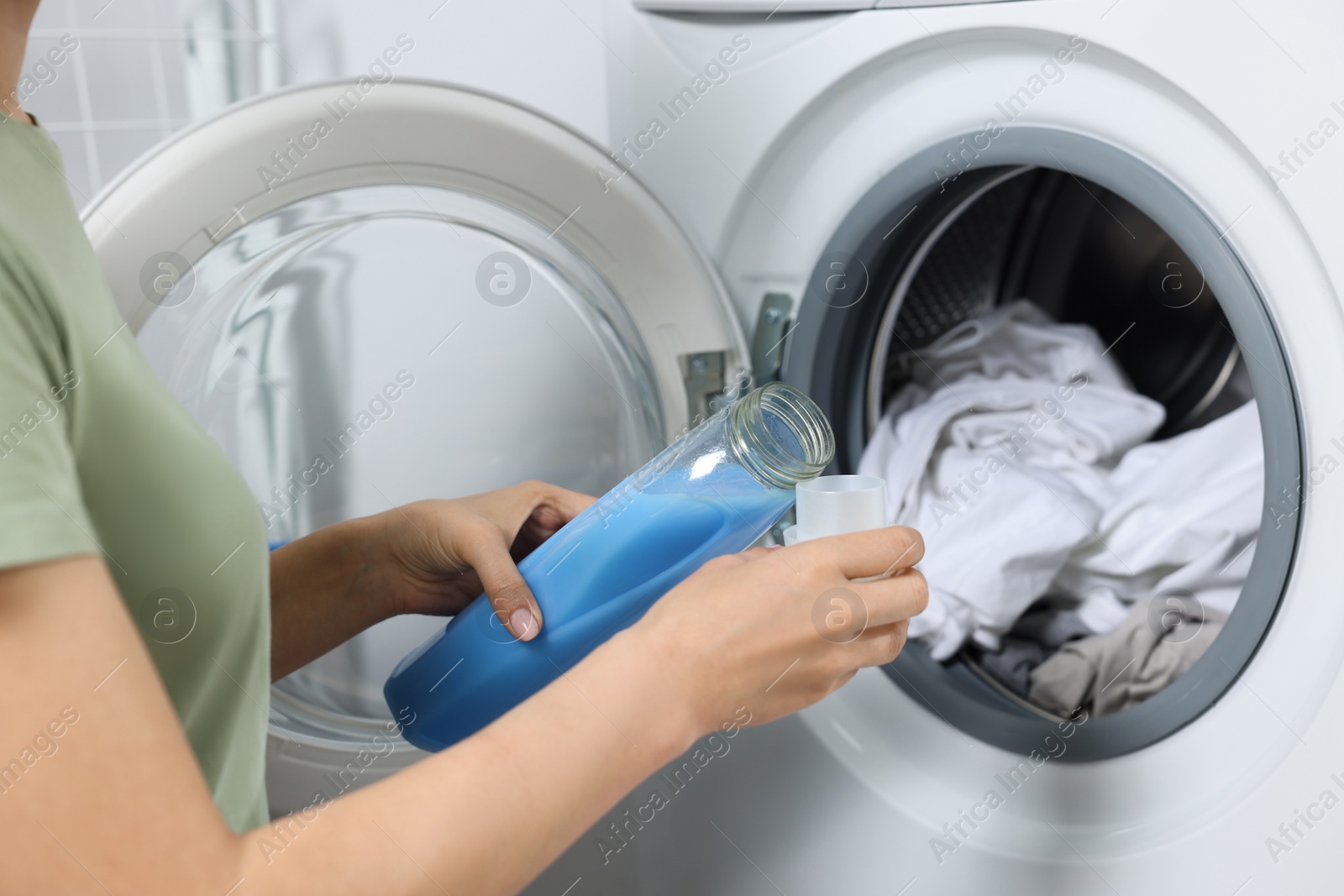 Photo of Woman pouring fabric softener into cap near washing machine with dirty clothes, closeup