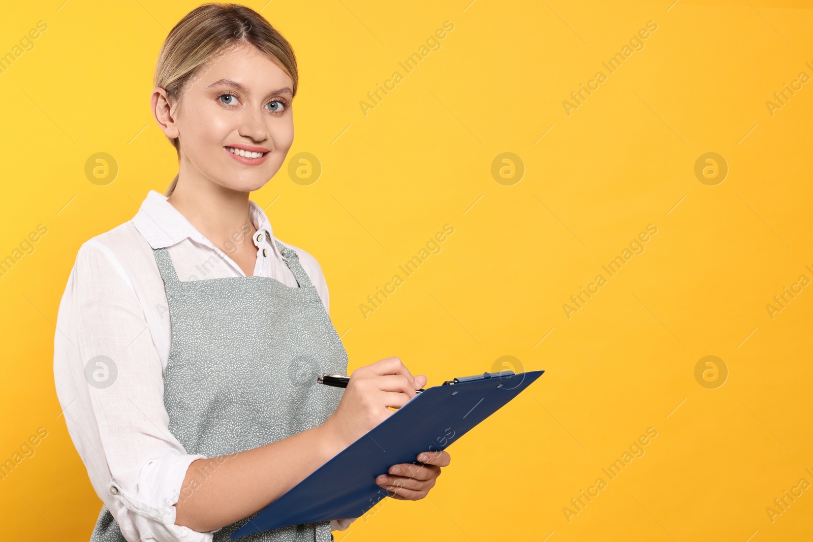 Photo of Beautiful young woman in clean apron with clipboard on orange background. Space for text