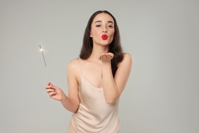 Christmas celebration. Beautiful young woman with sparkler blowing kiss on grey background