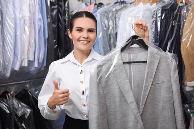 Dry-cleaning service. Happy worker holding hanger with jacket and showing thumb up indoors