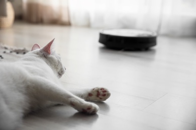 Photo of Cute white cat in room and modern robotic vacuum cleaner on background