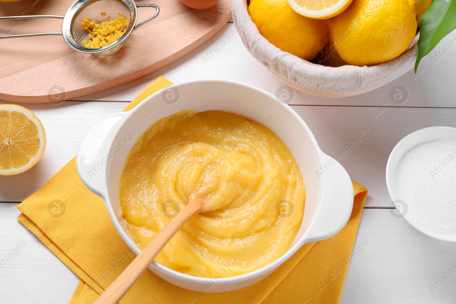 Photo of Delicious lemon curd in bowl, ingredients, spoon and sieve on white wooden table, flat lay