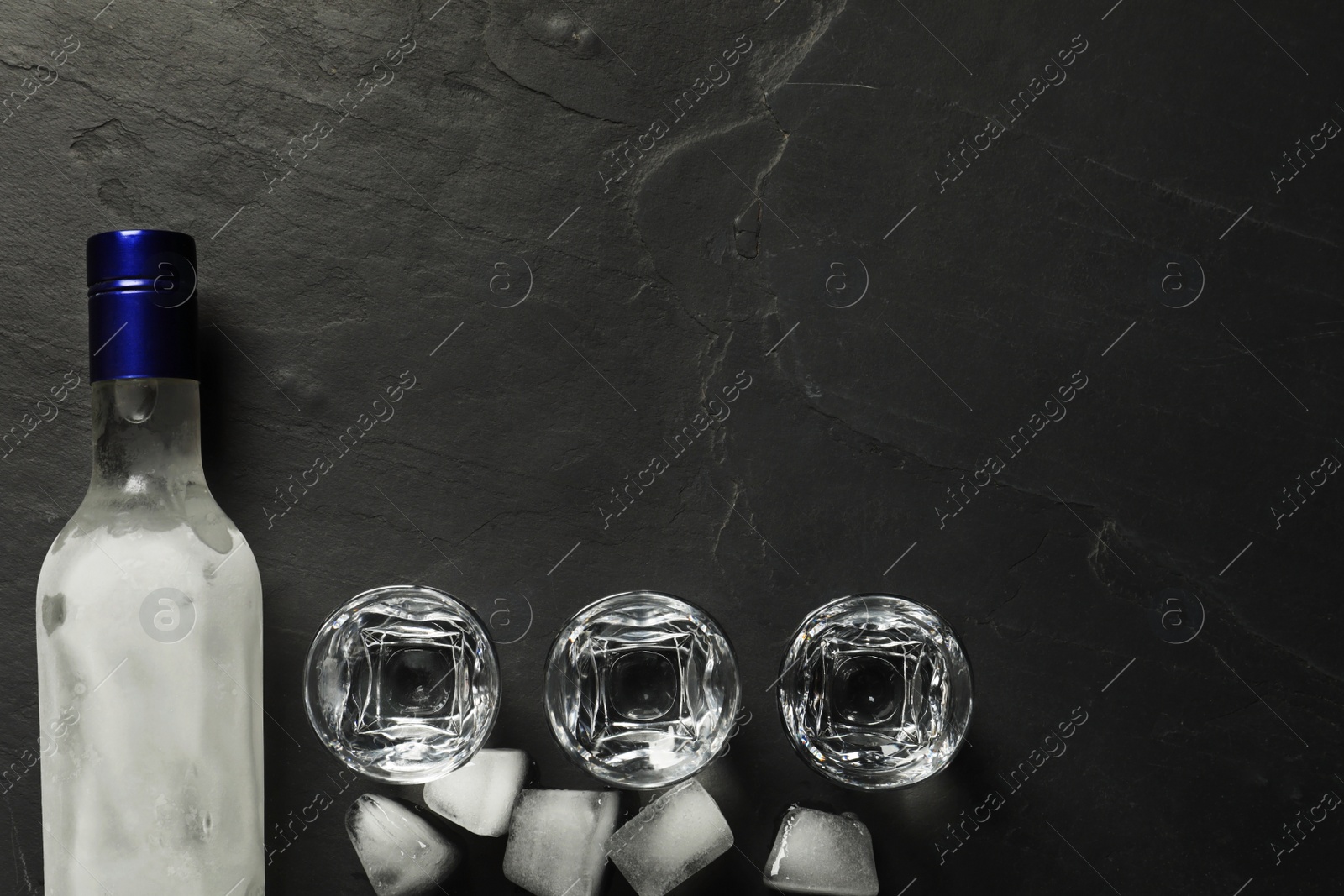 Photo of Bottle of vodka, shot glasses and ice cubes on black table, flat lay. Space for text