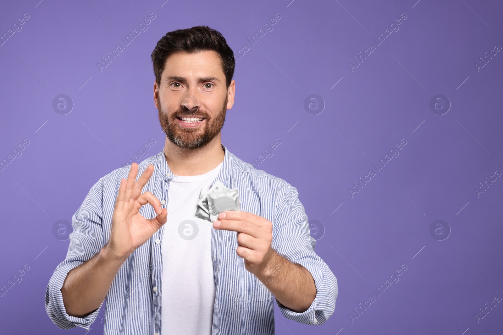 Photo of Man with condoms showing ok gesture on purple background, space for text. Safe sex