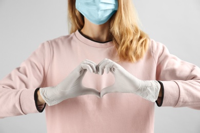 Photo of Young woman in medical gloves and protective mask making heart with hands on grey background, closeup