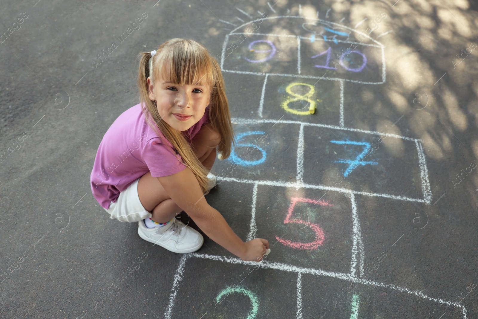 Photo of Little girl drawing hopscotch with chalk on asphalt outdoors. Happy childhood