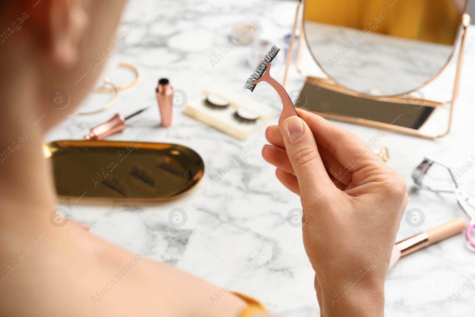 Photo of Woman holding tweezers with magnetic eyelashes at white marble table, closeup