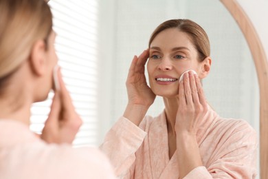 Photo of Beautiful woman removing makeup with cotton pad near mirror indoors