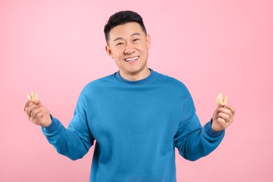 Photo of Happy asian man holding tasty fortune cookies with predictions on pink background