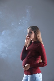 Young pregnant woman smoking cigarette on grey background. Harm to unborn baby