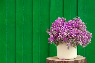 Beautiful lilac flowers in vase on wooden stump against green background, space for text