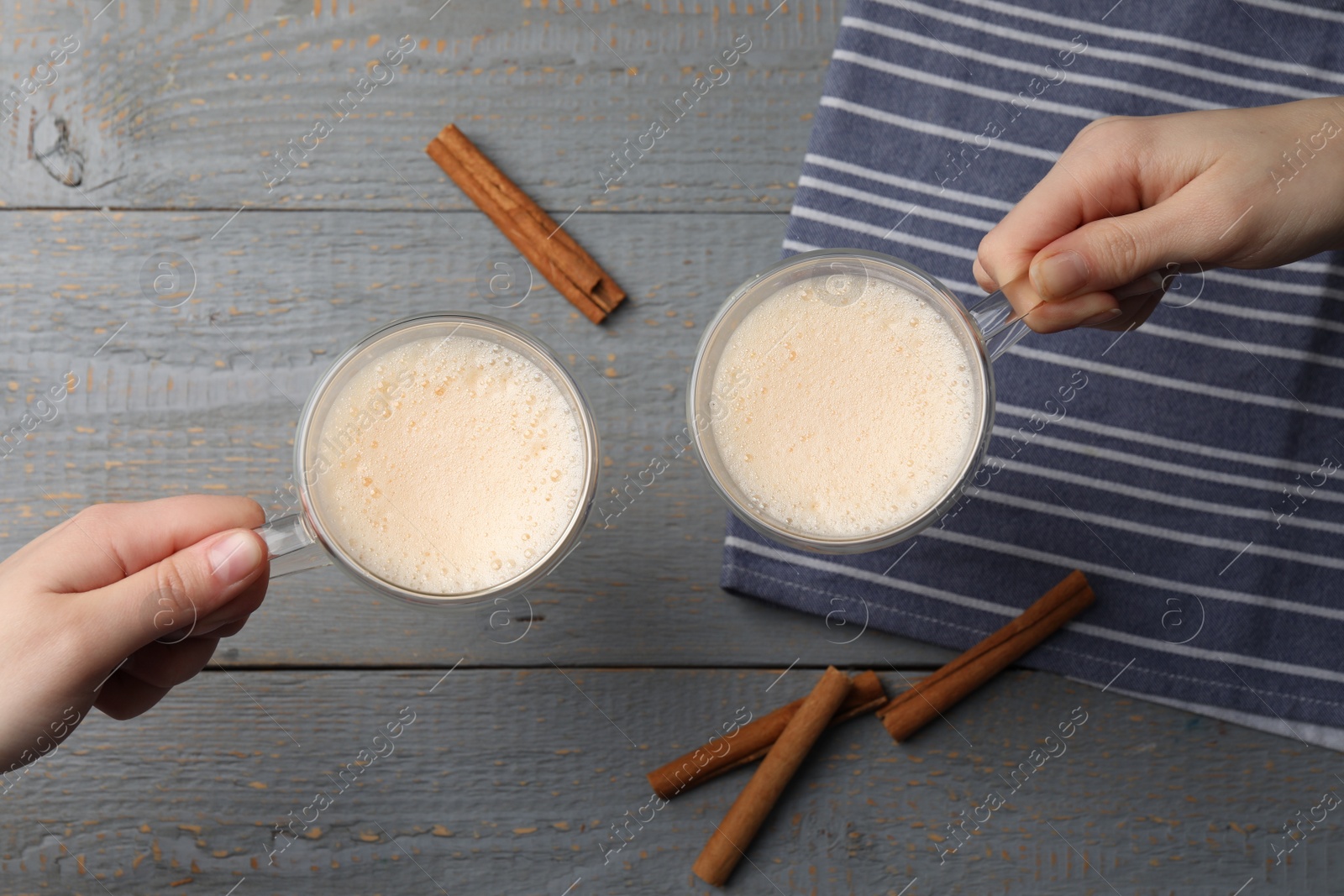 Photo of Women holding glass cups of delicious eggnog with cinnamon at grey wooden table, top view