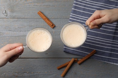 Women holding glass cups of delicious eggnog with cinnamon at grey wooden table, top view