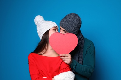 Photo of Young couple in warm clothes hiding behind heart while kissing on color background. Christmas celebration