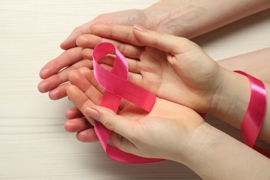 Women holding pink ribbon at white wooden table, closeup. Breast cancer awareness