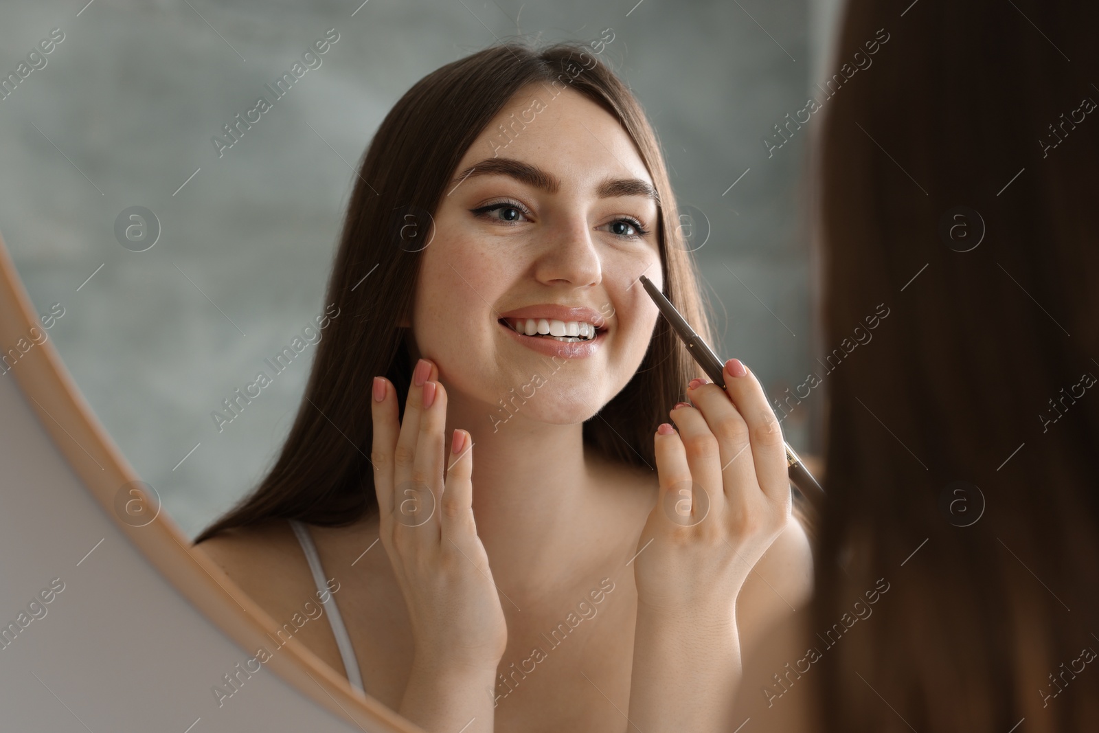 Photo of Smiling woman drawing freckles with pen near mirror indoors