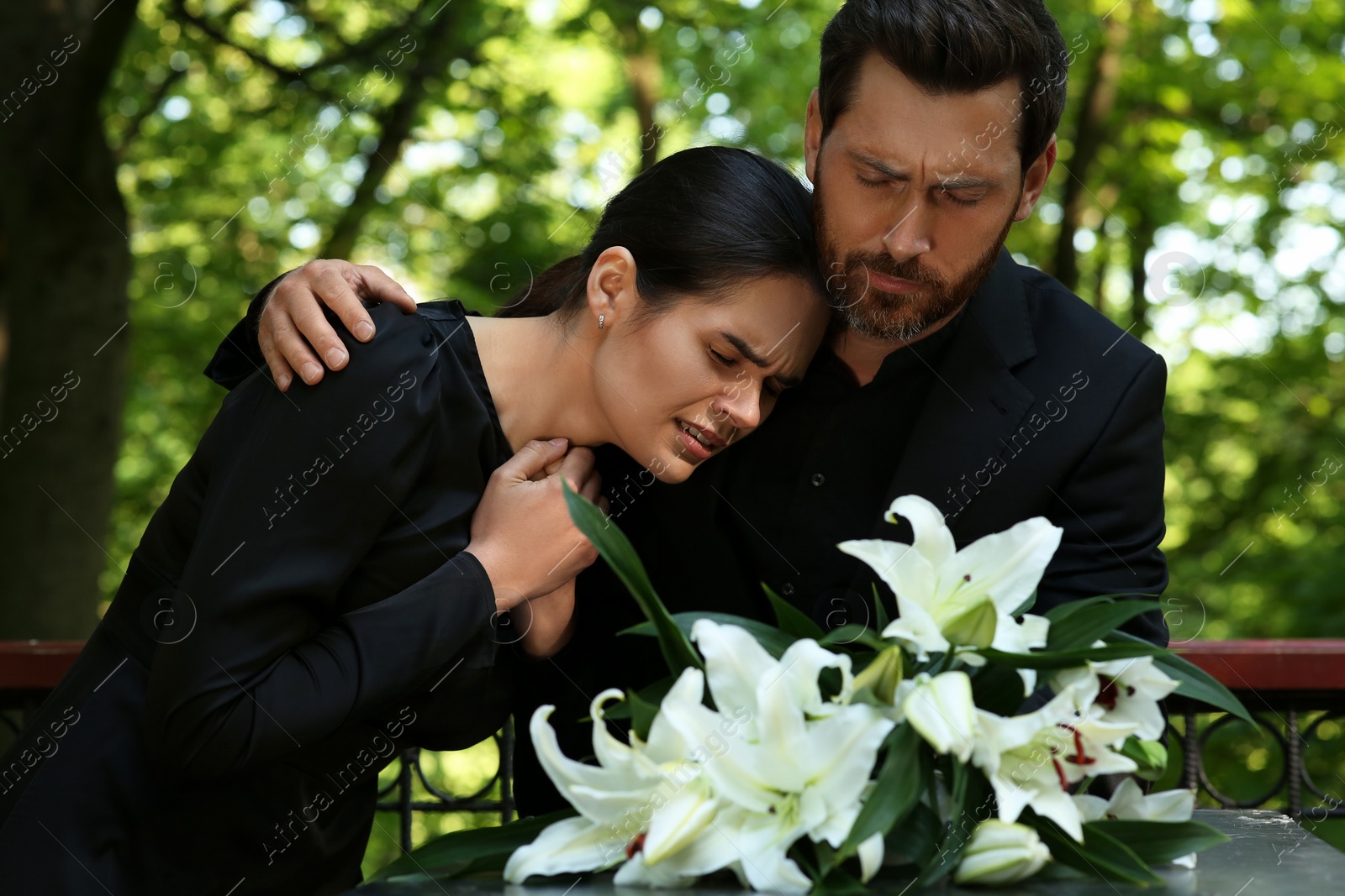 Photo of Sad couple mourning near granite tombstone with white lilies at cemetery. Funeral ceremony