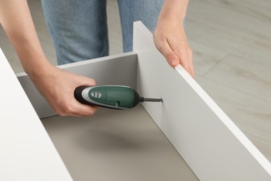 Photo of Woman with electric screwdriver assembling drawer indoors, closeup