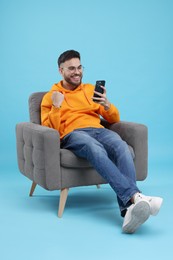 Happy young man using smartphone on armchair against light blue background