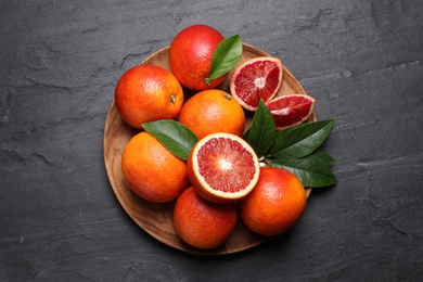 Photo of Plate of ripe red oranges and green leaves on dark table, top view