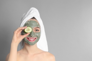 Photo of Young woman with clay mask on her face holding cucumber slice against grey background, space for text. Skin care