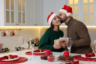 Photo of Happy young man in Santa hat presenting Christmas gift to his girlfriend at table in kitchen