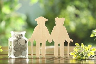 Pension savings. Figure of senior couple, coins in glass jar and green twig on wooden table outdoors