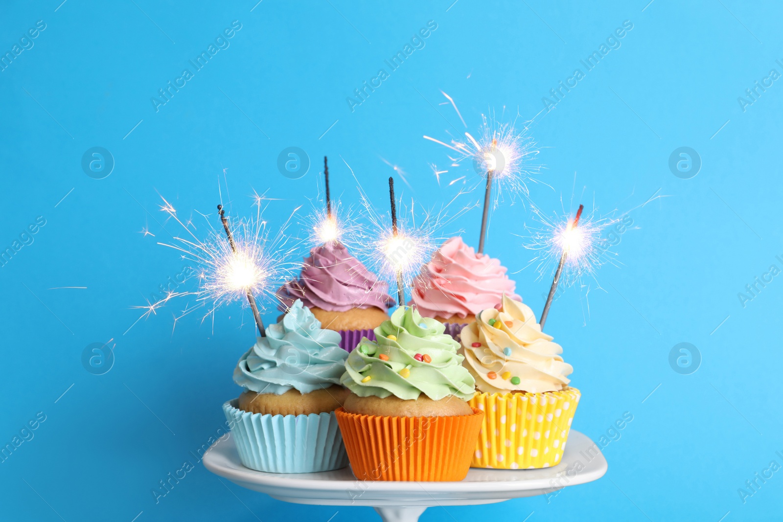 Photo of Birthday cupcakes with burning sparklers on stand against light blue background