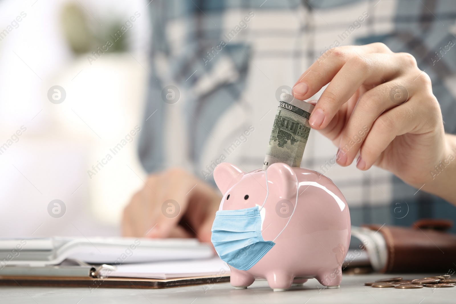 Image of Woman putting money into piggy bank with face mask at table indoors, closeup