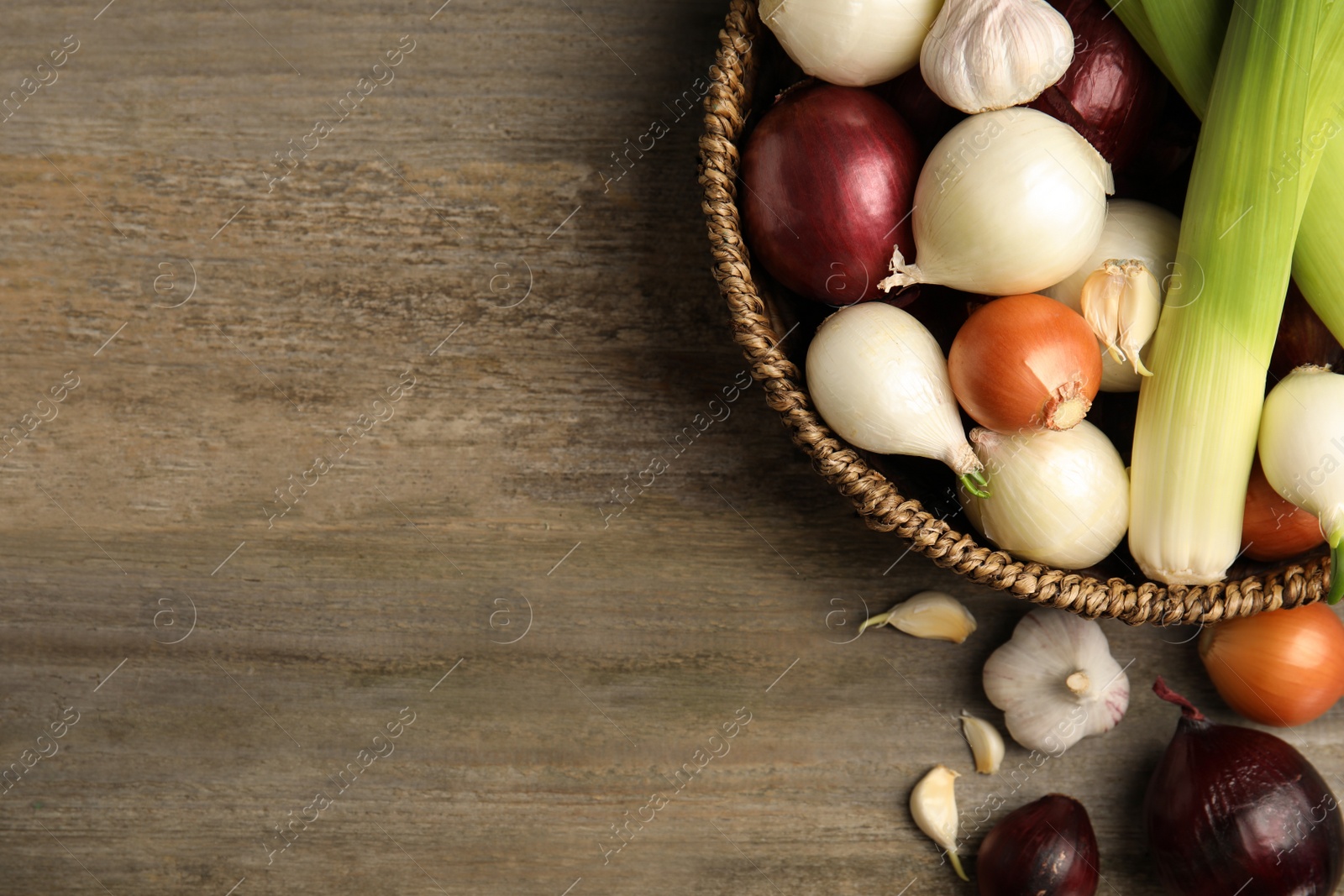 Photo of Wicker basket with fresh onion bulbs, leeks and garlic on wooden table, flat lay. Space for text