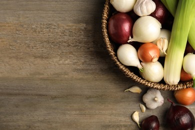 Wicker basket with fresh onion bulbs, leeks and garlic on wooden table, flat lay. Space for text