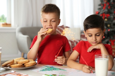 Photo of Little boy eating cookies while his friend drawing picture at home. Children celebrating Christmas