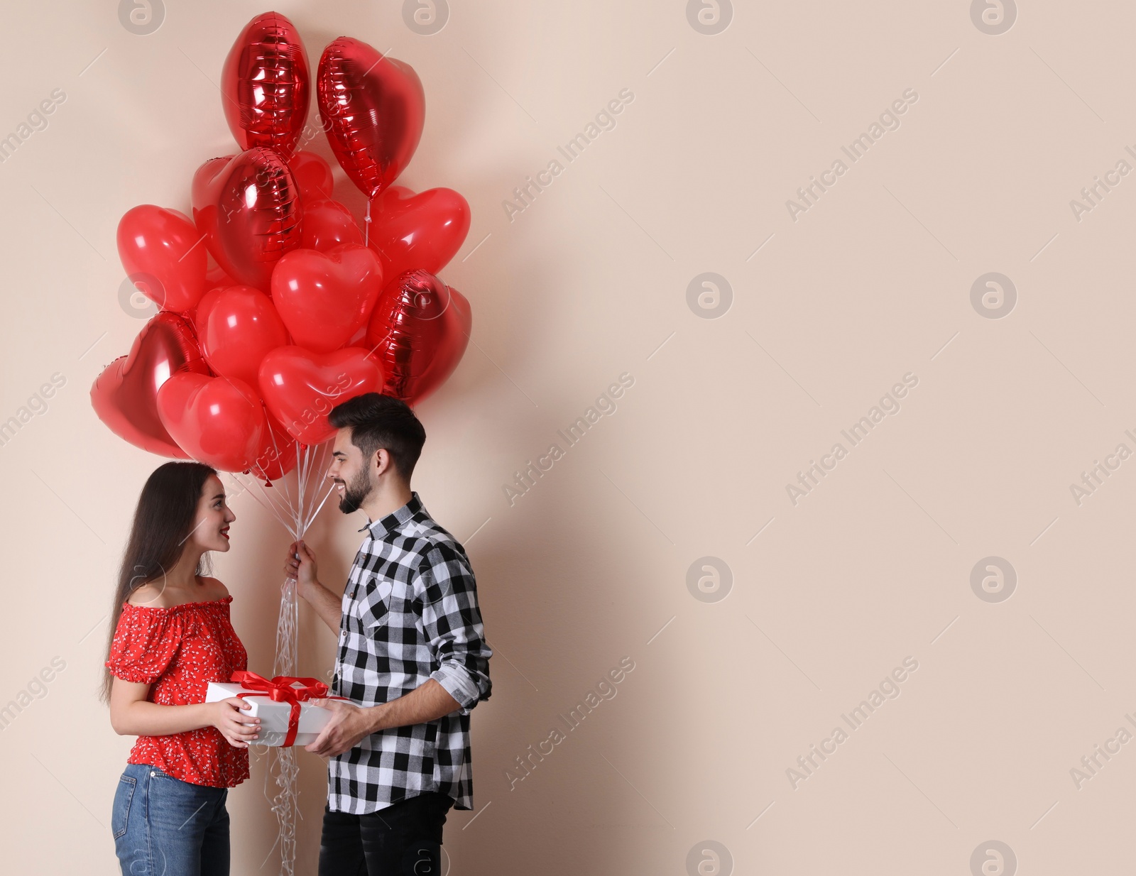 Photo of Happy young couple with gift box and heart shaped balloons on beige background. Valentine's day celebration