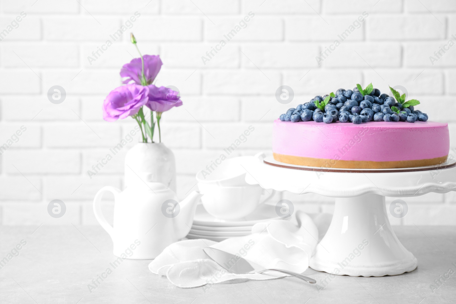 Photo of Stand with tasty blueberry cake and flowers on grey table against white brick wall. Space for text